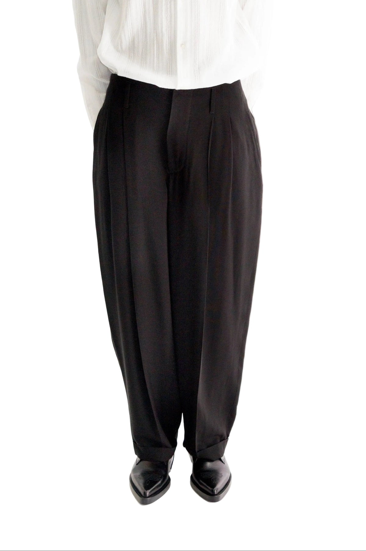 A person wearing black wide-leg pleated trousers with a white pleated Shirt and black pointed shoes, shown from the waist down against a white background.