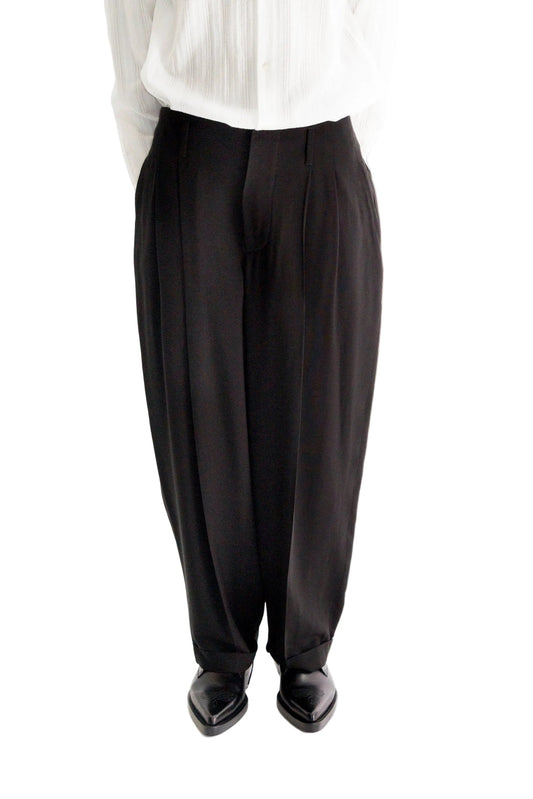 A person wearing black wide-leg pleated trousers with a white pleated Shirt and black pointed shoes, shown from the waist down against a white background.